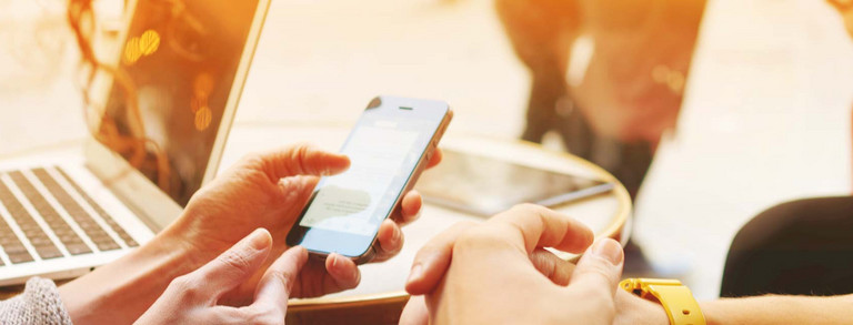 News Zwei Hände halten Mobiltelefon, im Hintergrund steht ein Laptop